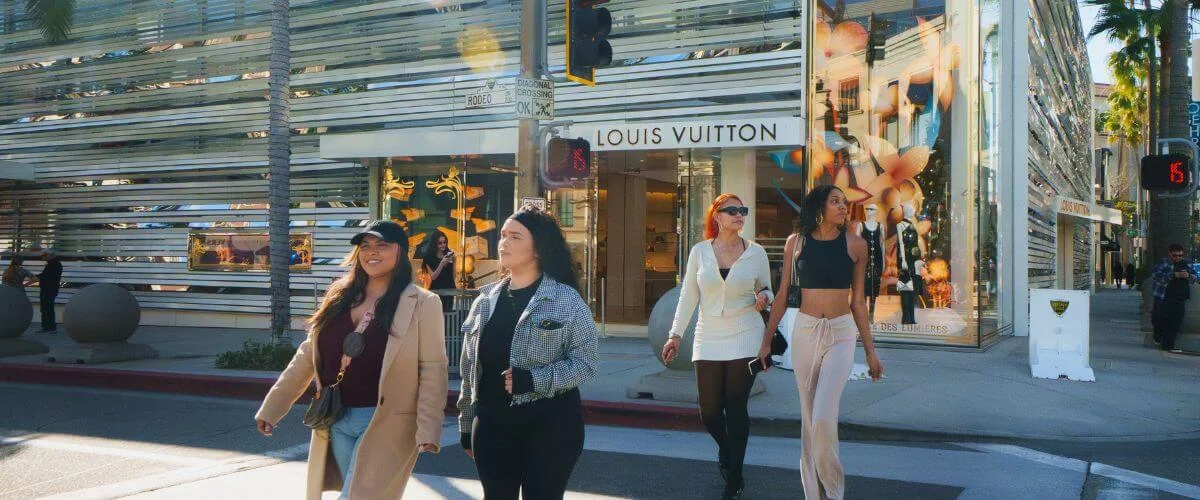 People walking past a Louis Vuitton store on Rodeo Drive in Beverly Hills, surrounded by palm trees and luxury storefronts on a sunny day.
