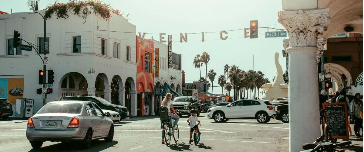 A busy street intersection in Venice Beach, Los Angeles, with a large "VENICE" sign stretching across Pacific Avenue. A woman and child cross on bicycles amid traffic, flanked by white arched storefronts, a colorful tattoo shop, and palm trees leading toward the beach.