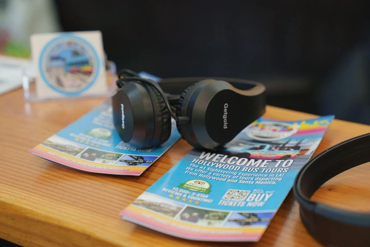 A pair of Getgold headphones rests on a colorful Hollywood Bus Tours welcome brochure on a wooden counter, advertising sightseeing tours departing from Hollywood and Santa Monica, with a tour logo card visible in the background.