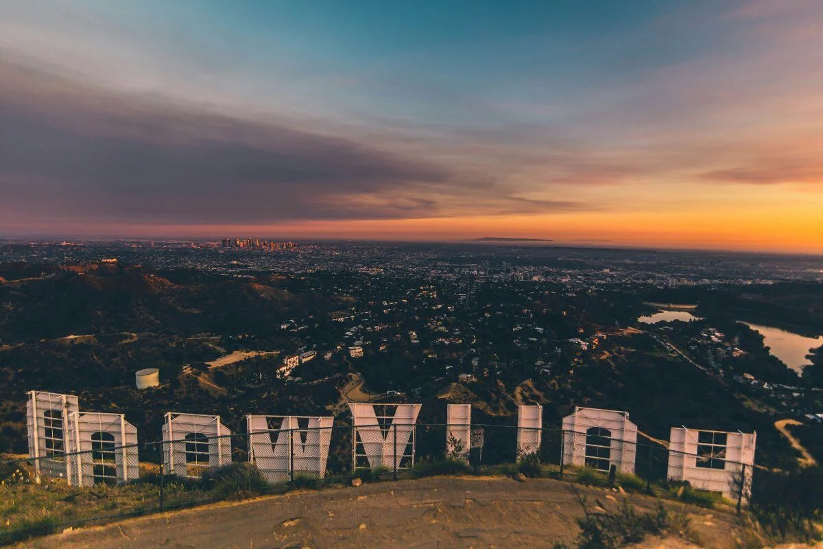 View from behind the Hollywood Sign looking over the vast sprawl of Los Angeles at sunset, with the back structure of the white letters visible in the foreground, the downtown skyline glowing in the distance, and a dramatic sky of orange, purple, and teal above.