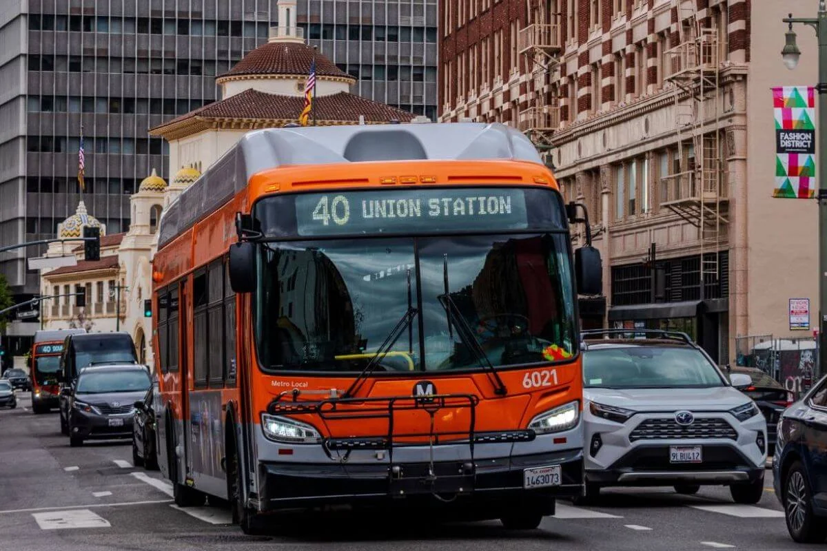 An orange LA Metro Local bus displaying Route 40 to Union Station drives through a busy downtown Los Angeles intersection, flanked by historic buildings, a domed structure, Fashion District banners, and surrounding traffic.