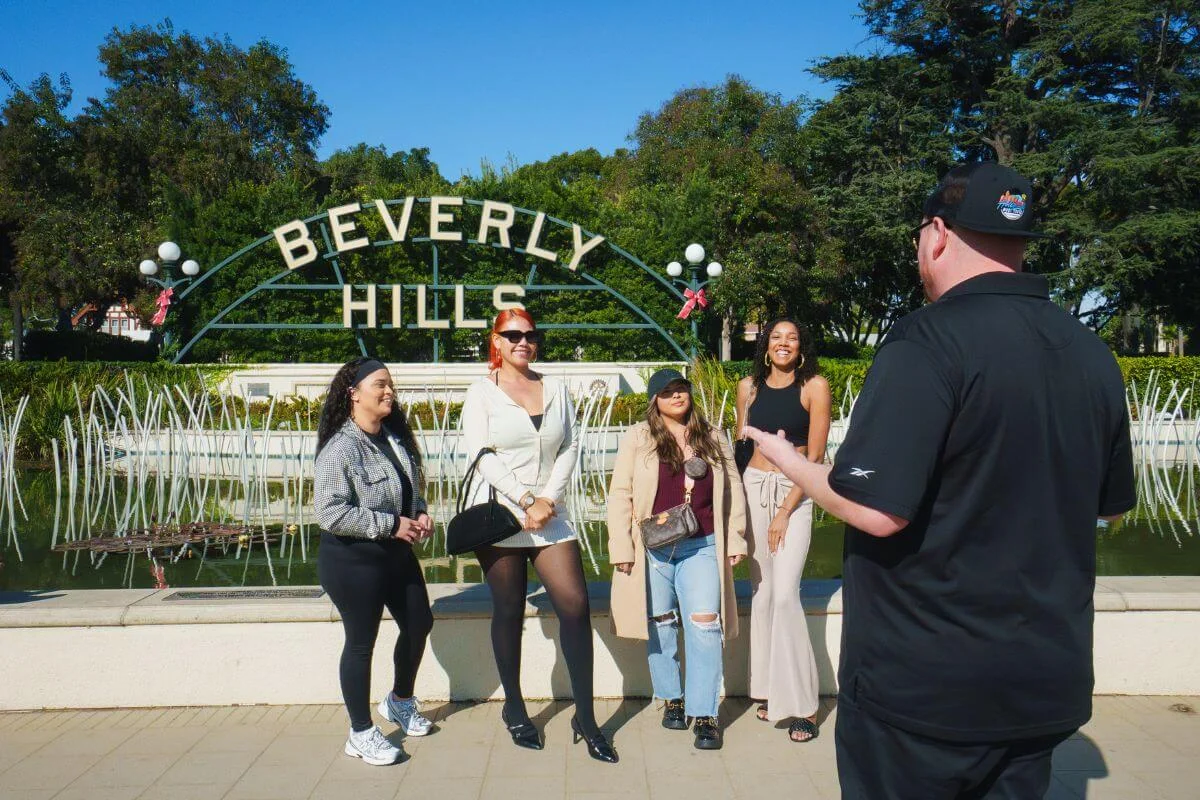 A tour guide in black speaks to four smiling women posing in front of the iconic green arch Beverly Hills sign, surrounded by manicured hedges, globe lamp posts, and tall trees under a clear blue sky.