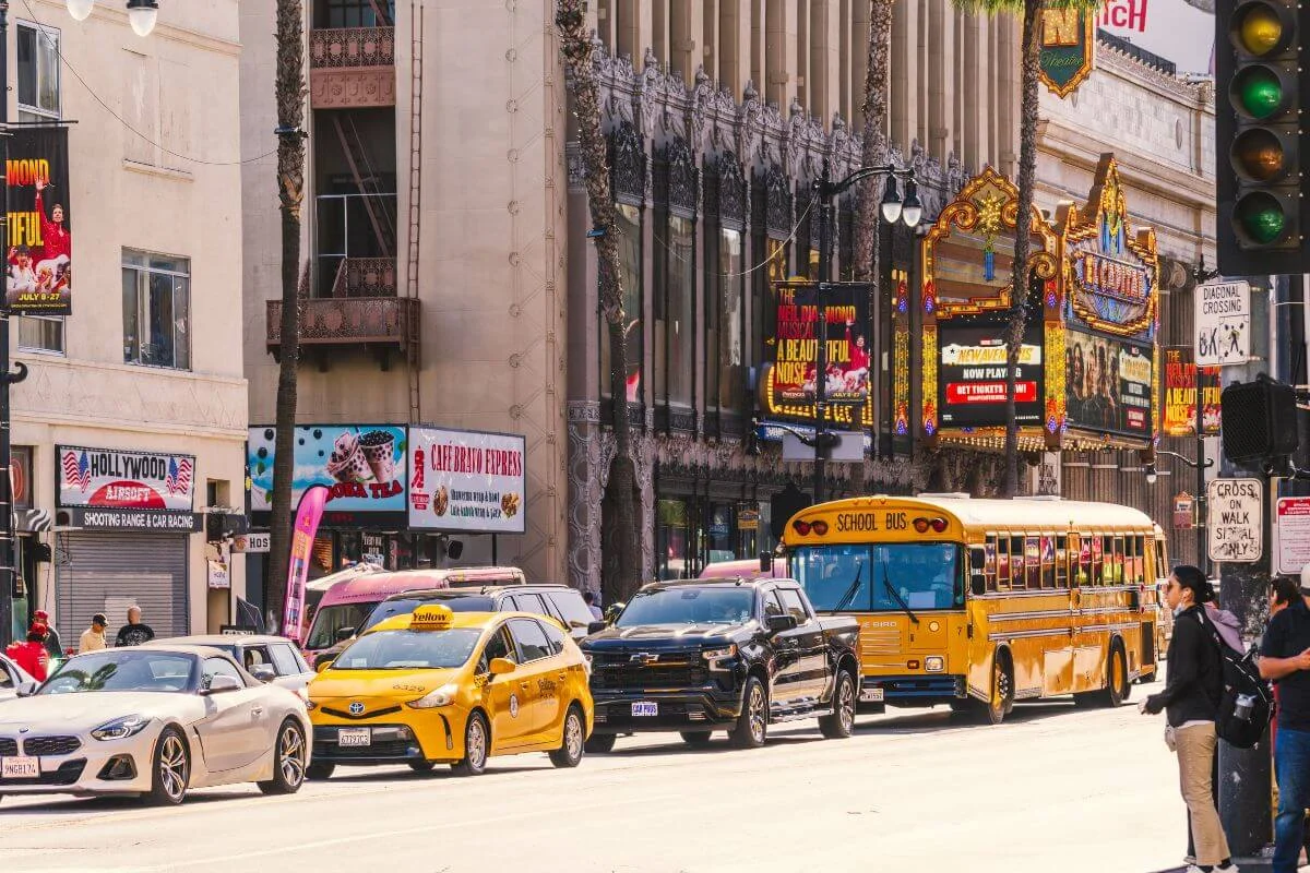 A congested Hollywood Boulevard street scene with a yellow taxi, school bus, and SUV in traffic, flanked by ornate theater marquees, digital billboards, and retail storefronts under bright midday sunshine.