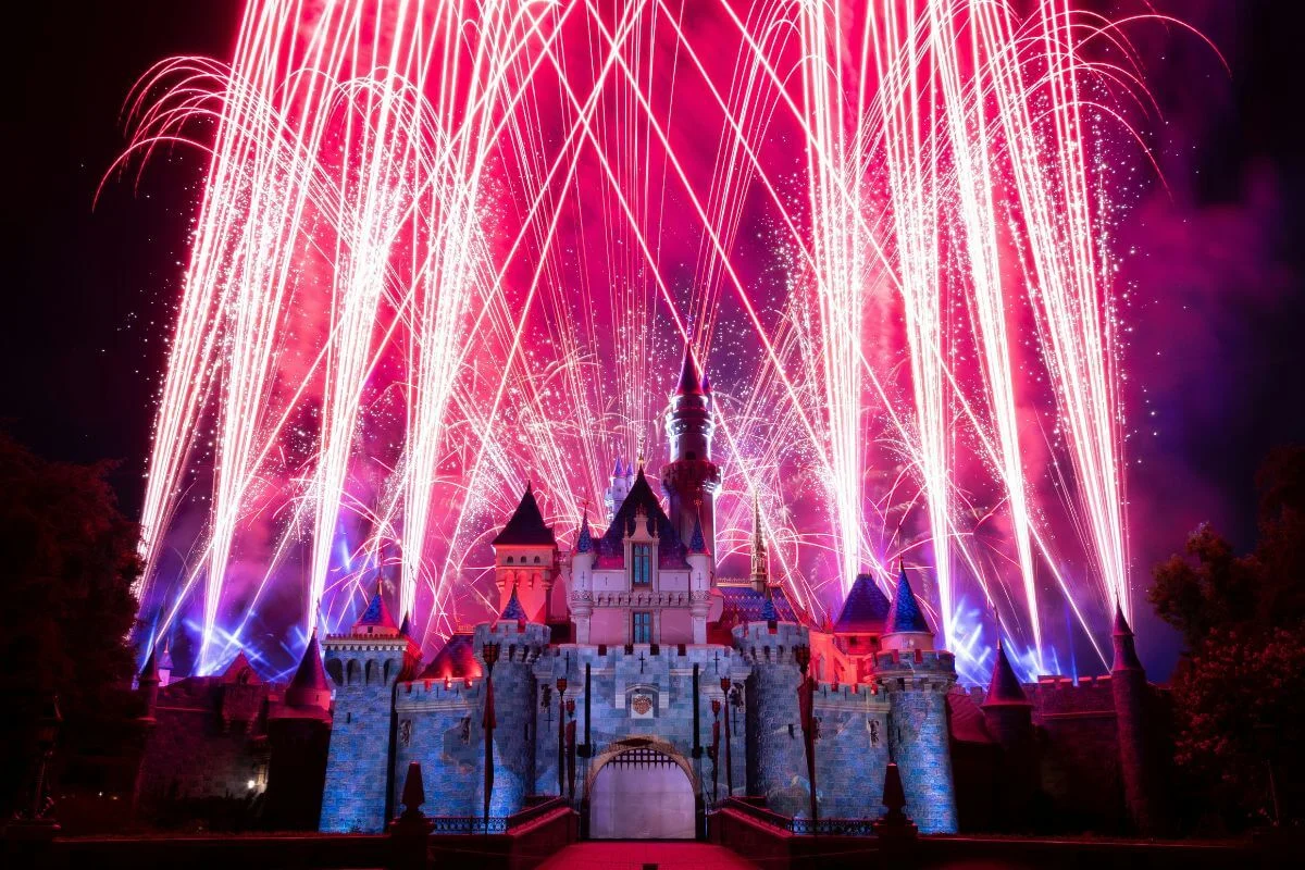 A spectacular nighttime fireworks display erupts in vivid pink, red, and white streaks above the illuminated Sleeping Beauty Castle at Disneyland, with blue projection lighting on the castle walls and a deep red sky above.