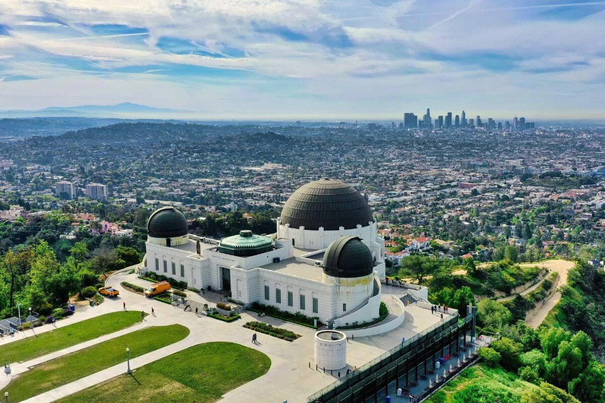 Aerial view of the Griffith Observatory in Los Angeles, showing its white Art Deco facade and three bronze domes on a hilltop, with manicured green lawns, school buses in the parking area, and the sprawling LA cityscape and downtown skyline stretching into the hazy distance under a partly cloudy sky.