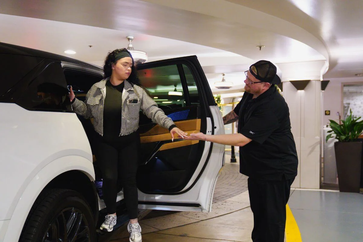 A hotel valet in all-black uniform reaches out to assist a young woman stepping out of a white luxury SUV with a wood-trimmed interior, inside a covered hotel driveway with warm overhead lighting.