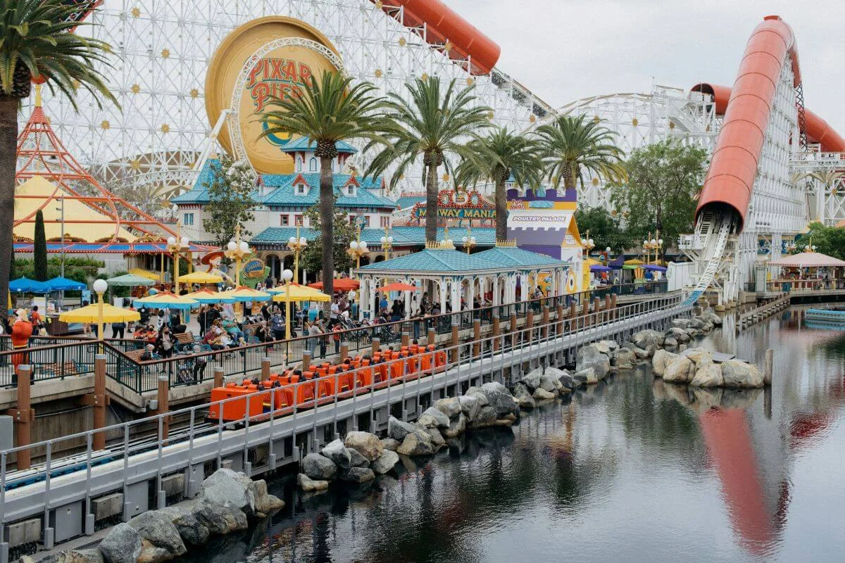 A wide view of Pixar Pier at Disney California Adventure, featuring the Incredicoaster roller coaster, Midway Mania attraction, colorful storefronts, palm trees, orange ride cars on a waterfront track, and guests dining under yellow umbrellas, reflected in the calm lagoon below.