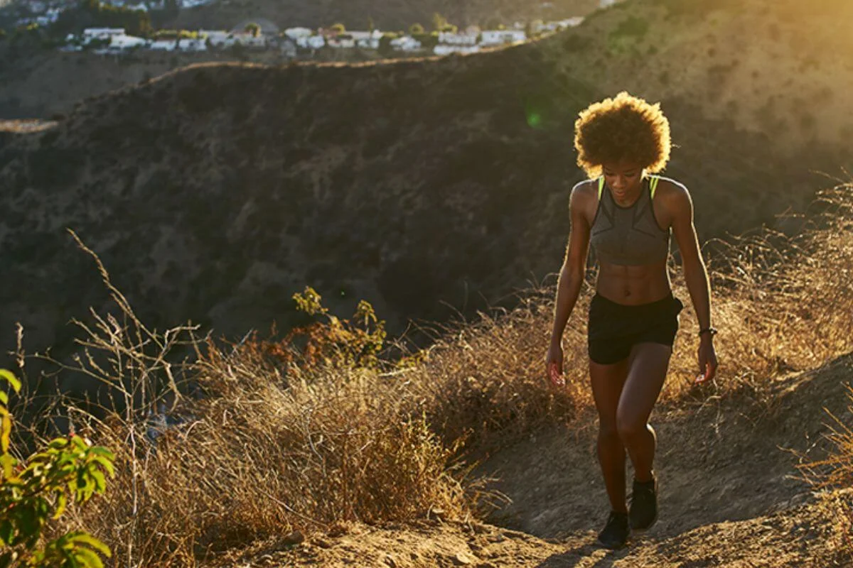 A fit woman in a gray crop top, black shorts, and sneakers hikes up a dry, sun-baked dirt trail on a Los Angeles hillside, with golden sunlight illuminating the dried grass and shrubs around her and residential buildings visible on the ridge behind her.