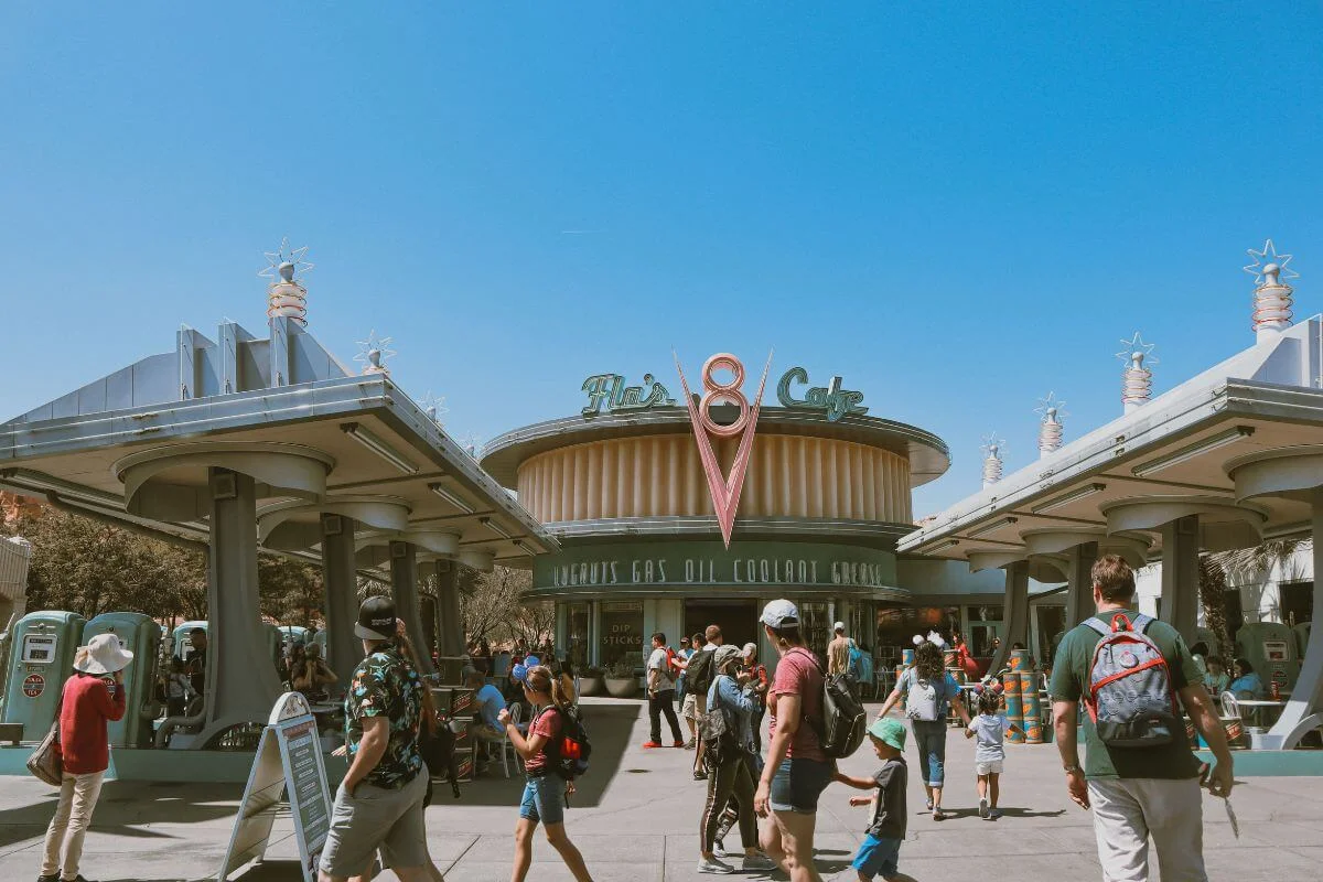 Crowds of park guests walk toward the retro-styled Flo's V8 Cafe in Cars Land at Disney California Adventure, a circular diner building with a large V8 logo and vintage gas pump decor under a bright blue sky.