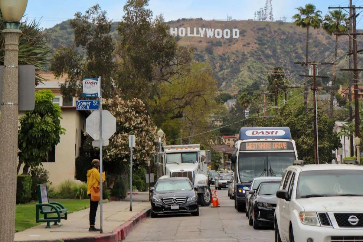 A DASH bus headed to Westshire Drive travels along Gower Street in Hollywood, with the iconic Hollywood Sign clearly visible on the brown hillside above the residential neighborhood, as a pedestrian in a yellow jacket waits at the corner.