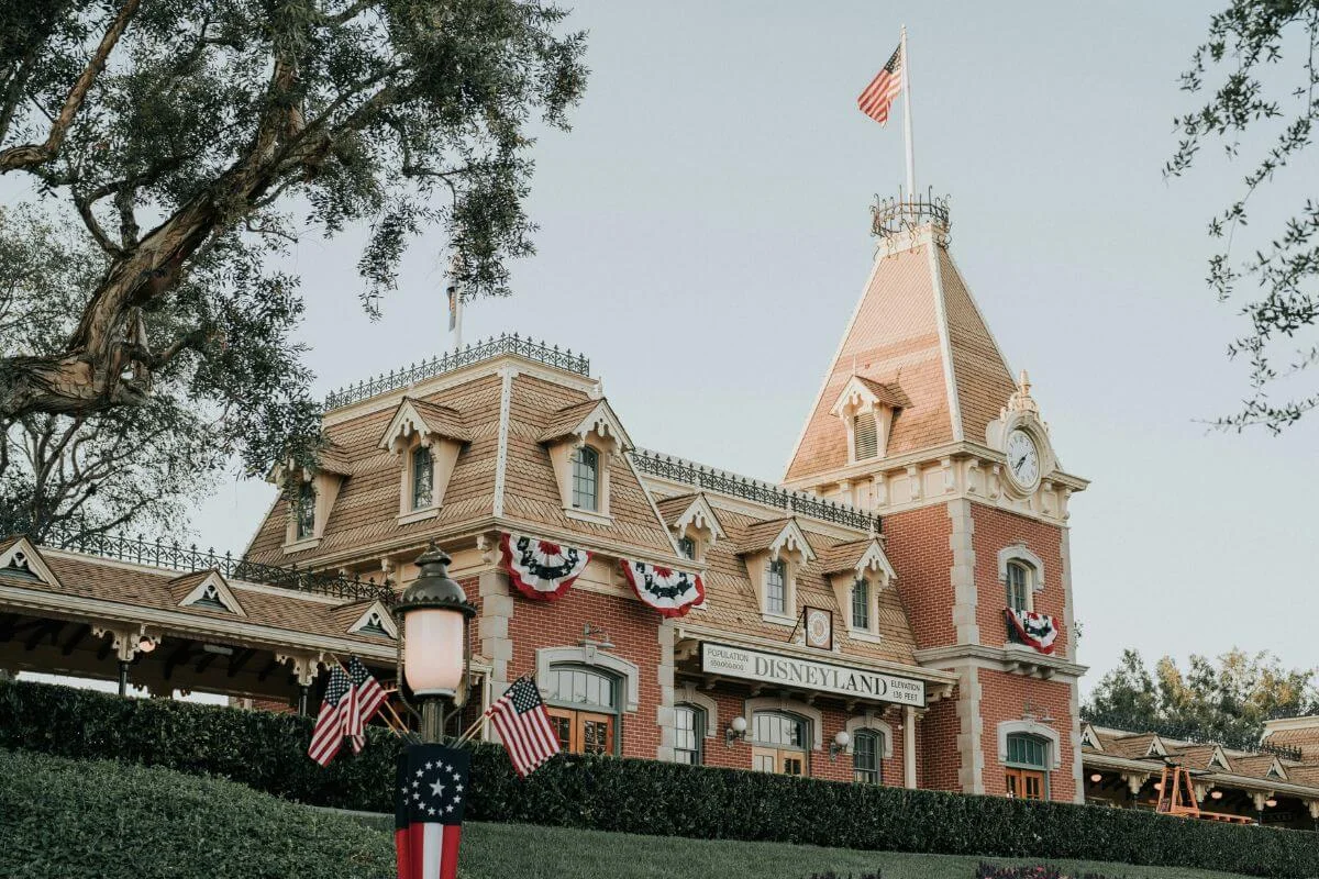 The iconic red brick Disneyland train station entrance building adorned with red, white, and blue patriotic bunting and American flags, with a clock tower, ornate roof details, and an American flag flying from the top under a soft overcast sky.