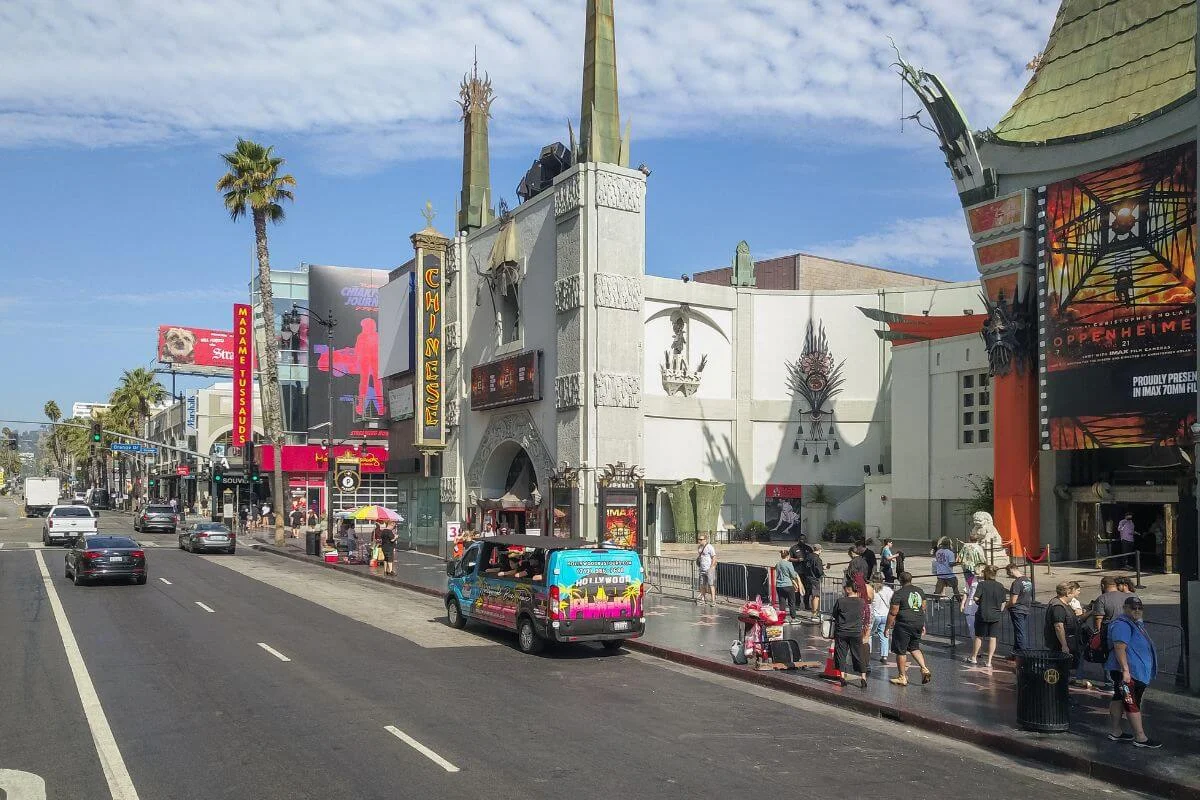 A daytime street view of the TCL Chinese Theatre and surrounding Hollywood Boulevard attractions, with tourists on the Walk of Fame, a colorful Hollywood tour van, Madame Tussauds signage, and an Oppenheimer movie poster on the right under a bright blue sky.