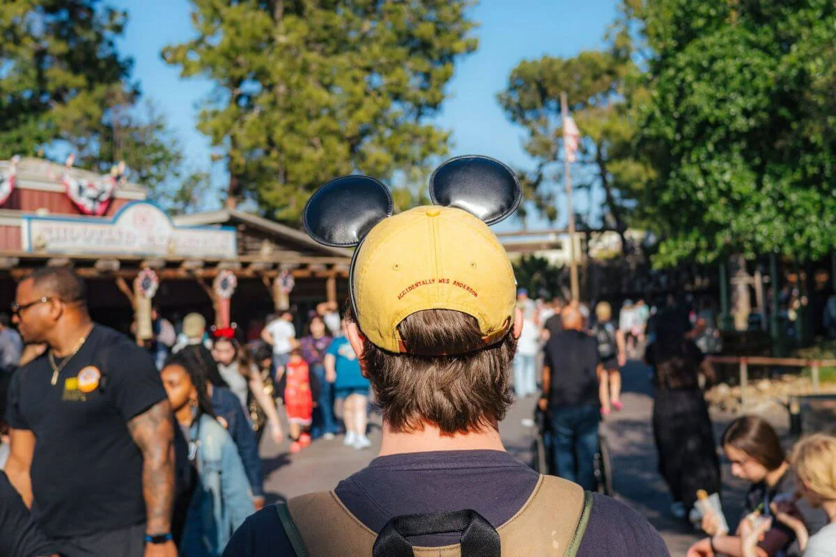 Back view of a visitor wearing a yellow baseball cap with black Mickey Mouse ears walking through a busy outdoor area of a theme park, with crowds of guests, rustic wooden buildings, and tall trees visible in the sunny background.