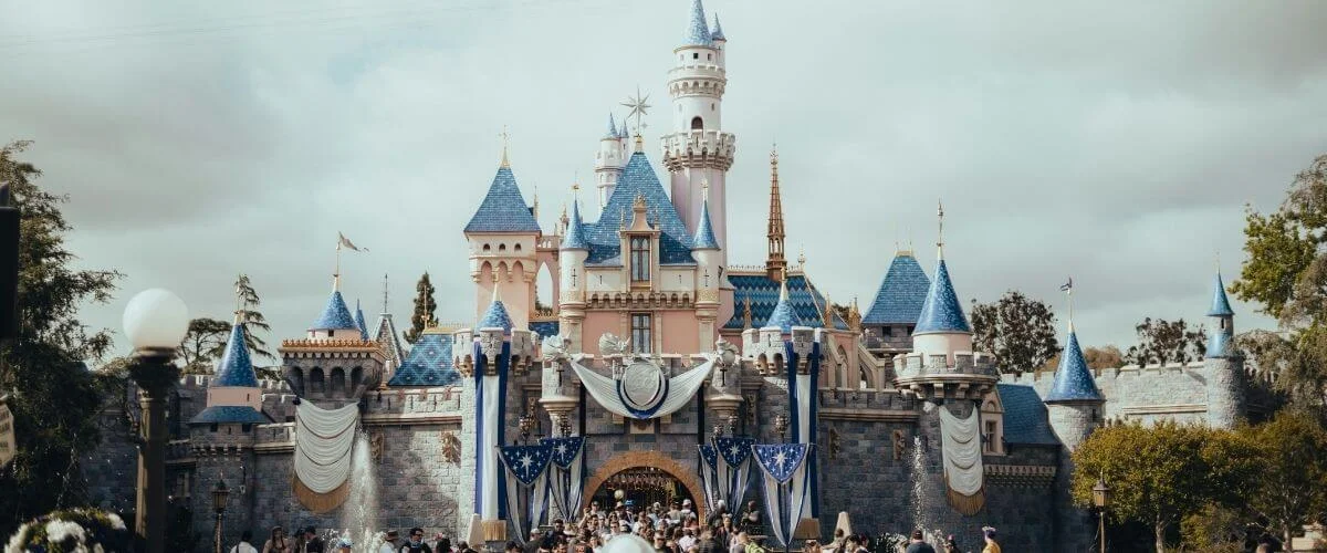 A front-facing view of Sleeping Beauty Castle at Disneyland in Anaheim, adorned with blue and white celebratory draping and fountains on either side, with crowds of park guests gathered at the entrance under a dramatic overcast sky.