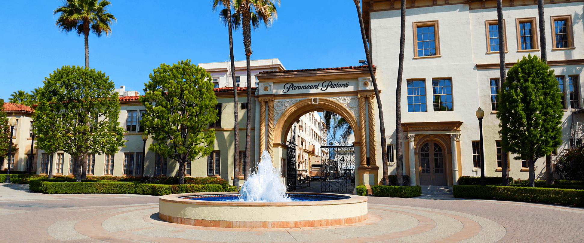 The iconic Paramount Pictures ornamental arch gate and circular fountain at the center of the studio lot, framed by tall palm trees and Spanish Colonial style buildings under a bright blue sky.