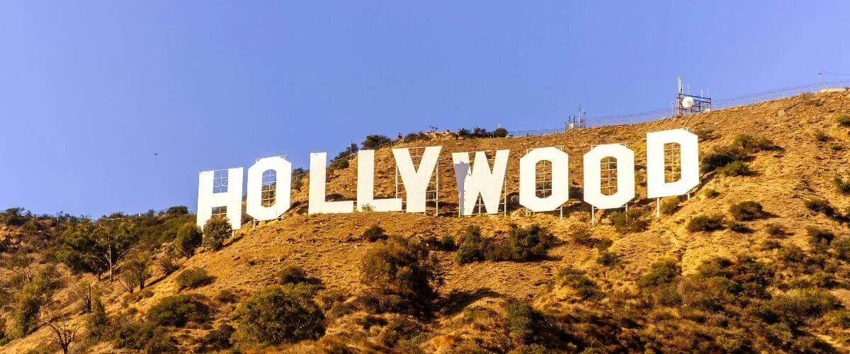 A straight-on view of the iconic white Hollywood Sign mounted on a dry, shrub-covered hillside under a bright clear blue sky, with small figures of visitors visible near the letters and a communications tower on the ridge above.