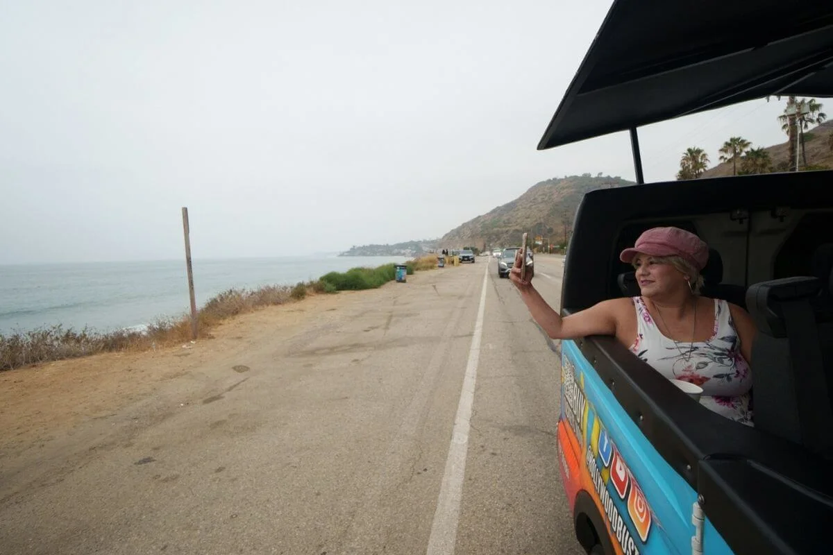 Woman leaning out of a Hollywood Bus Tours open-air van taking a photo along the PCH coastal highway