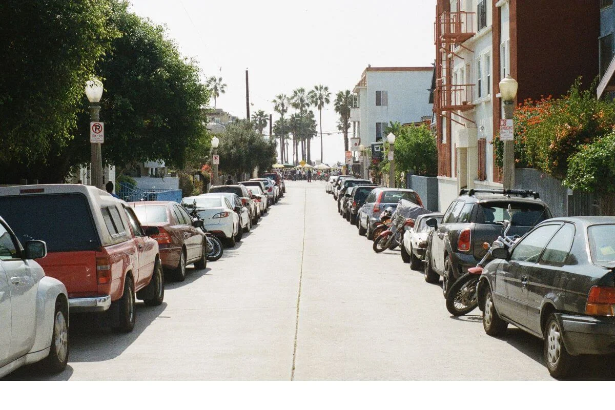 Narrow street lined with parked cars and palm trees leading toward a beachside neighborhood