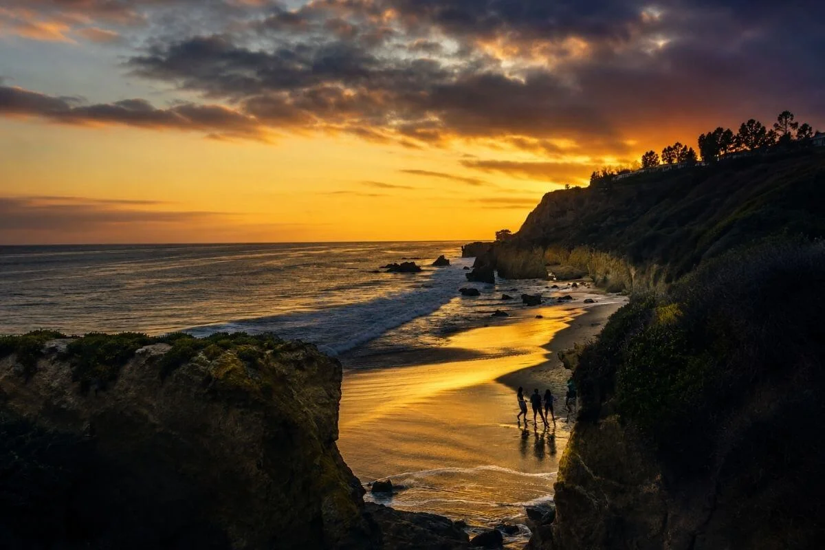 Dramatic orange sunset over rocky cliffs and shoreline with silhouettes of people walking on the beach