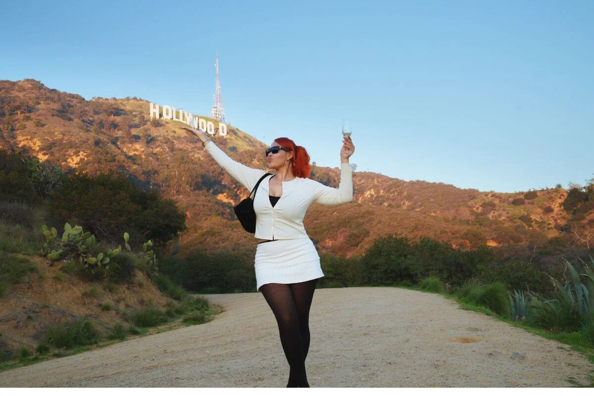 Woman in white outfit holding a wine glass and posing on a dirt trail with the Hollywood Sign behind her