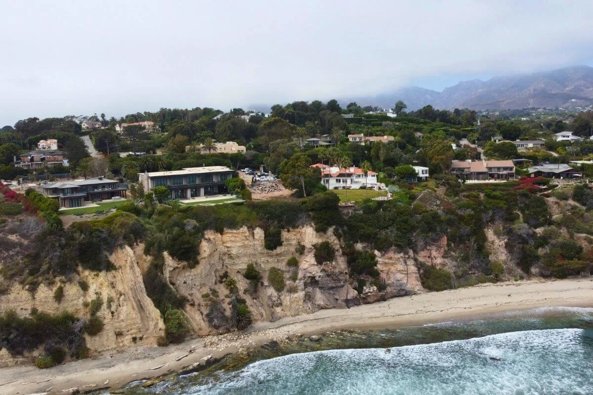 Drone view of upscale homes perched on sandy cliffs above a Malibu beach with ocean waves below