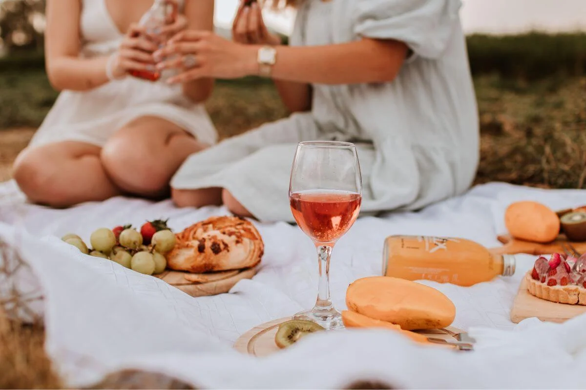 Glass of rose wine beside grapes, croissant, and fruit on a white blanket with two women sitting behind