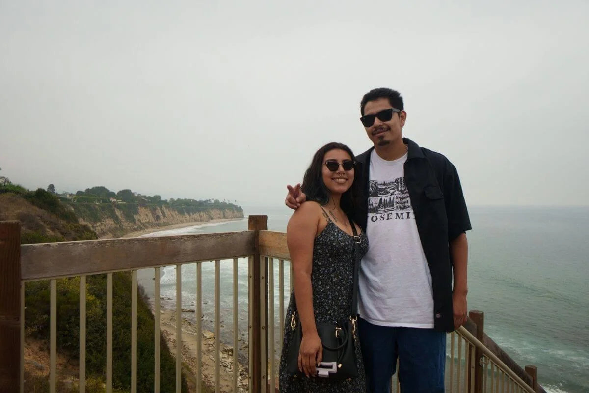 Couple smiling at a clifftop railing with Malibu's rocky coastline and ocean stretching behind them