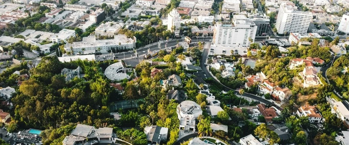 Aerial view of Los Angeles at golden hour, showing hillside homes with pools and palm trees in the foreground and the flat city grid stretching toward the horizon.