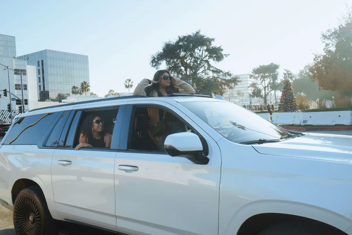 A woman looks out the passenger window while another stands through the open sunroof of a white SUV parked along a city street.