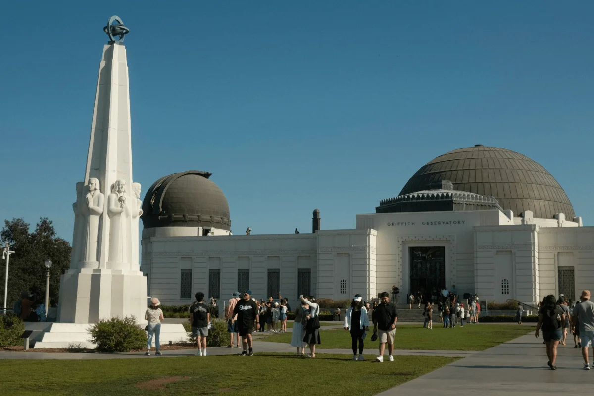 Visitors walking across the grassy front lawn in front of the white Griffith Observatory building and Astronomers Monument under a clear blue sky
