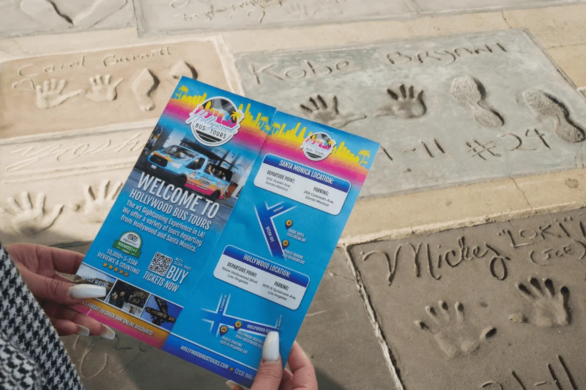 A traveler holding a colorful Hollywood Bus Tours flyer over handprints and signatures embedded in concrete outside the TCL Chinese Theatre in Hollywood