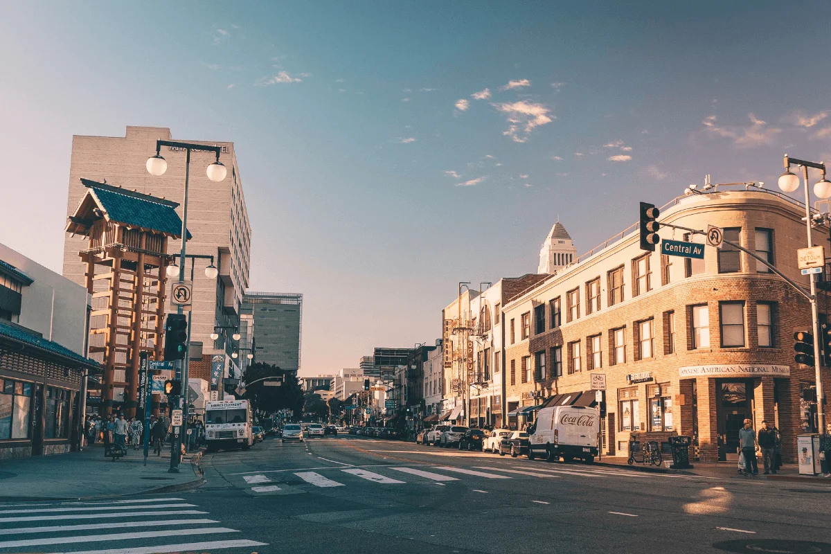 A wide street view of First Street in Little Tokyo, Los Angeles, showing the traditional red Yagura fire tower on the left, local storefronts, pedestrians, and City Hall in the background under a warm sky.