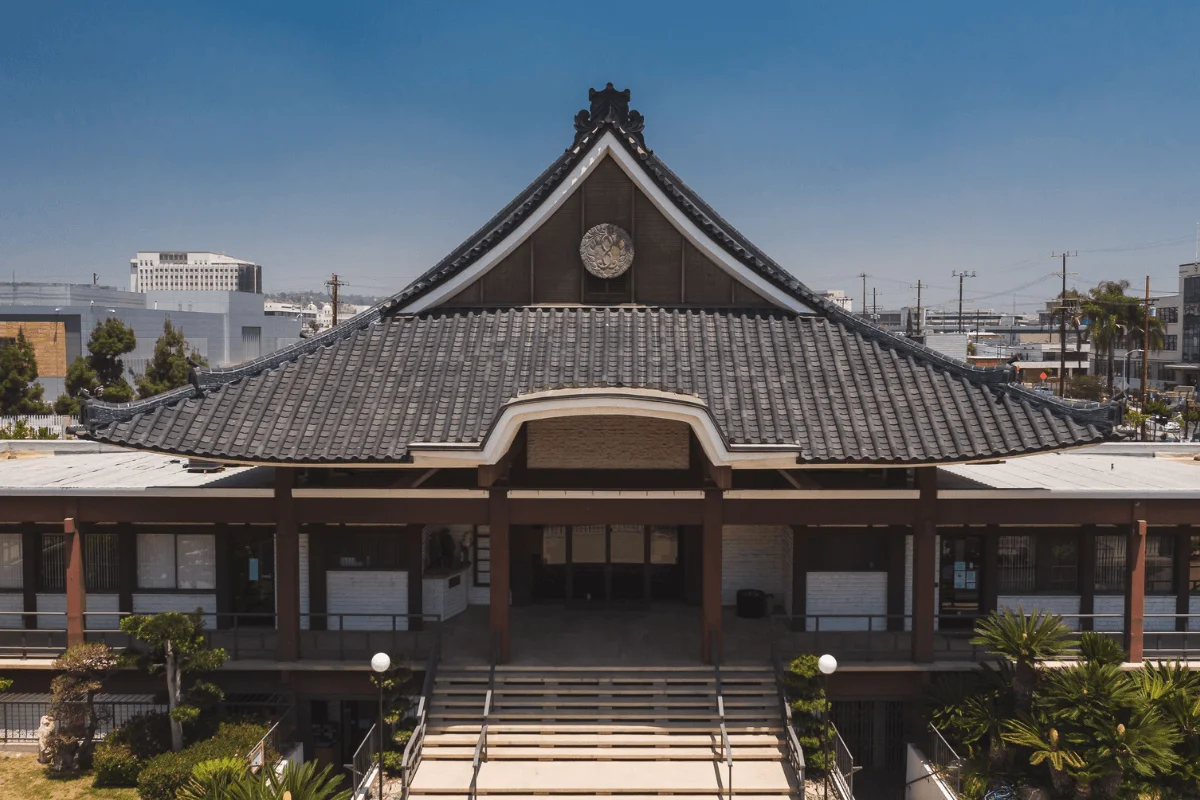 A front-facing shot of the historic Koyasan Buddhist Temple in Little Tokyo, showcasing its traditional dark tiled roof, wide front staircase, and surrounding green palm trees under a bright blue sky.