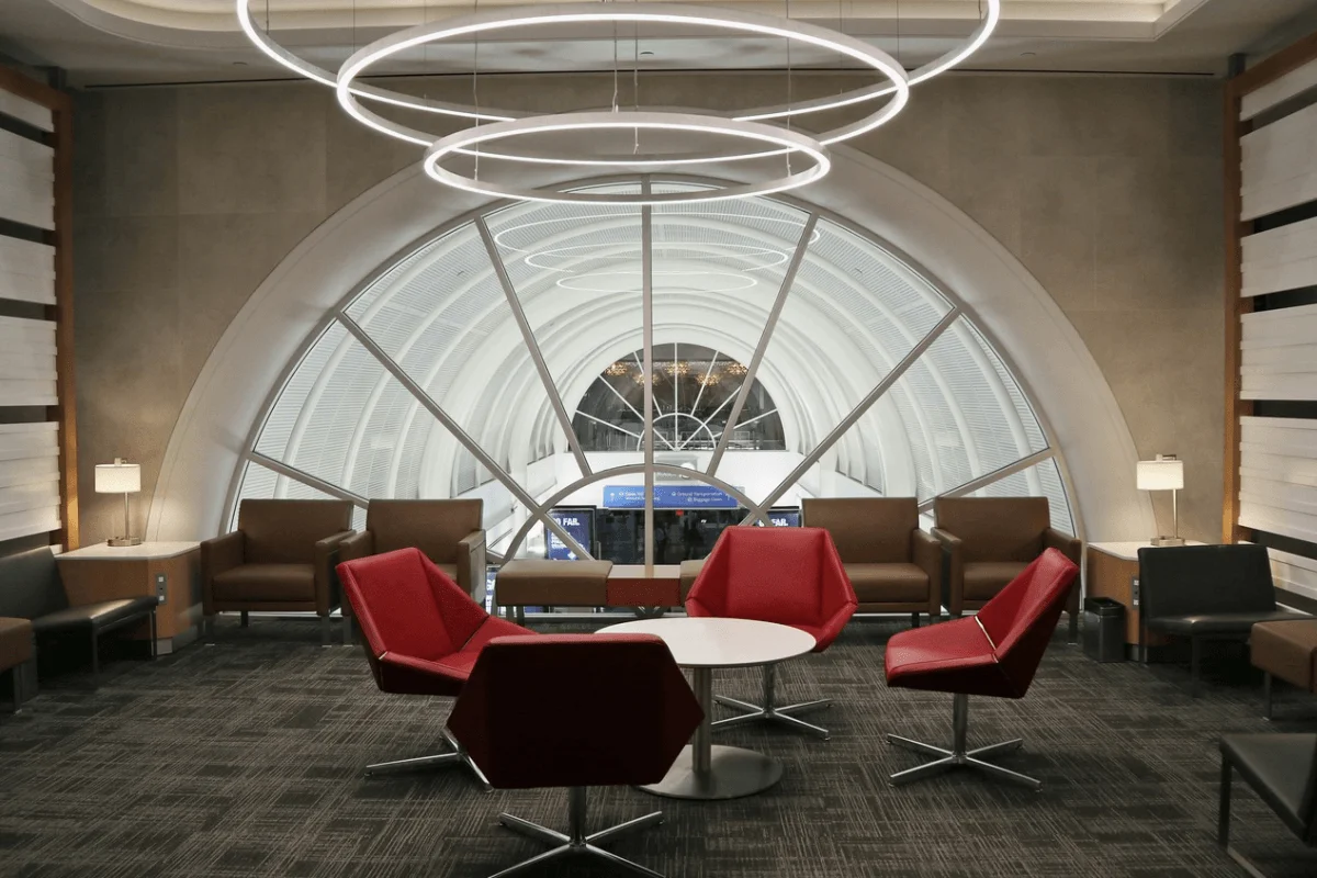 A quiet, modern airport terminal lounge featuring red and brown armchairs positioned around a small coffee table in front of a large arched window.