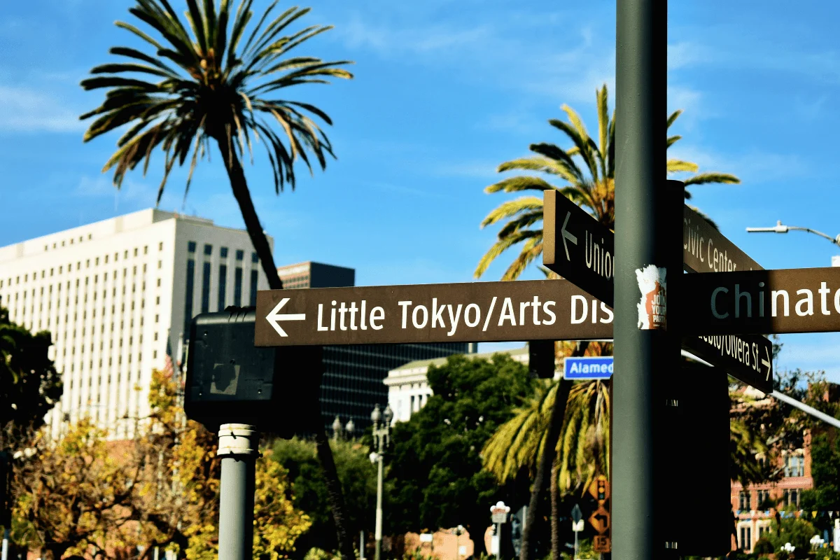 A brown directional street sign in Los Angeles pointing left toward Little Tokyo and the Arts District, framed by tall green palm trees against a clear blue sky.