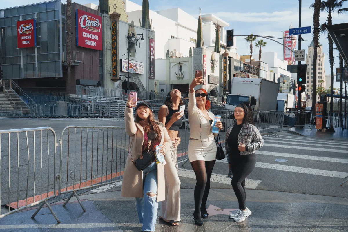 A group of four young female tourists taking selfies with smartphones while standing on the Hollywood Walk of Fame crosswalk, with TCL Chinese Theatre visible in the background.