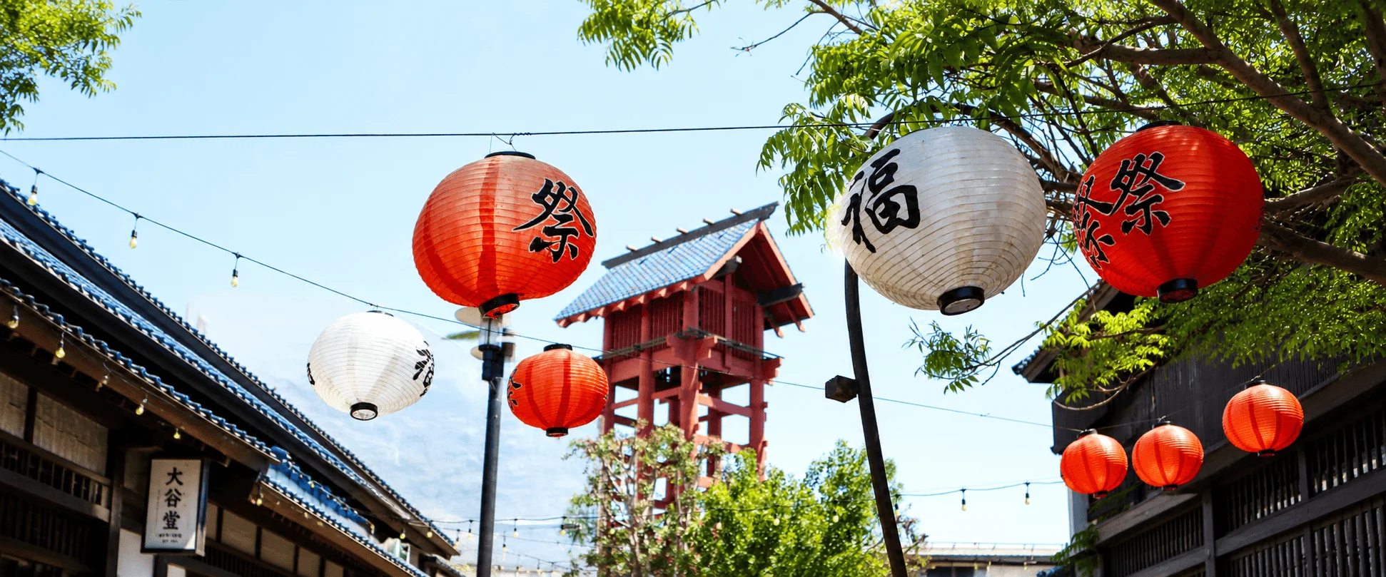 Red and white paper lanterns with Japanese kanji characters hanging overhead along a pedestrian walkway in Japanese Village Plaza, with traditional blue-tiled rooftops and a red wooden tower in the background.