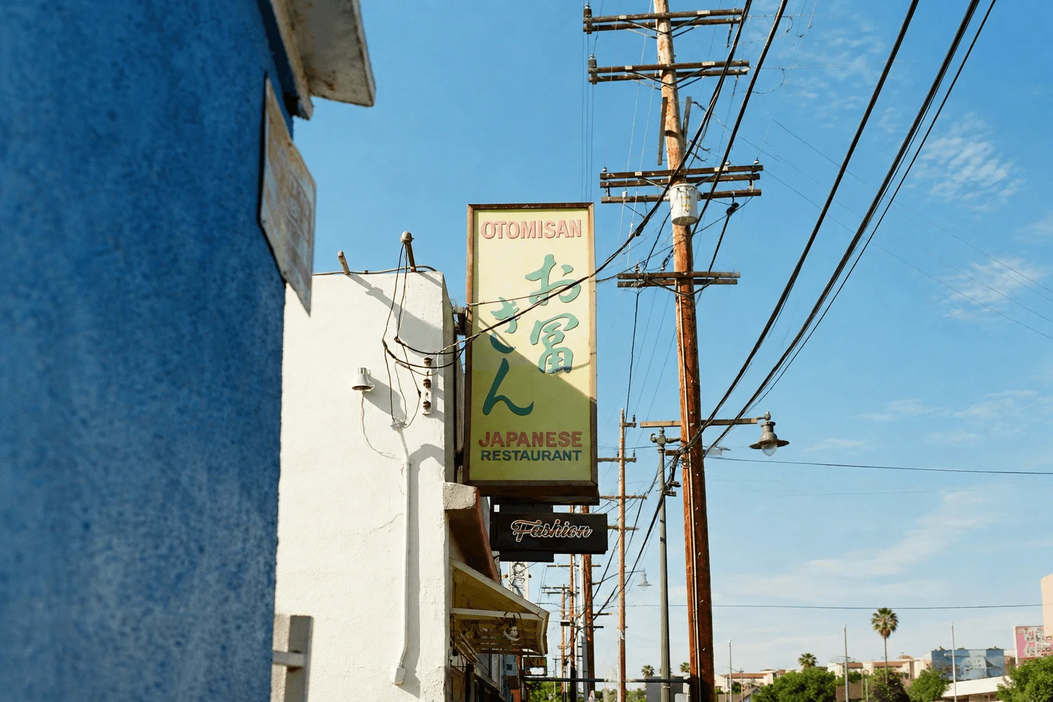 A retro vertical sign for Otomisan Japanese Restaurant featuring Japanese characters, mounted on a white building against a bright blue sky with wooden power poles and wires.