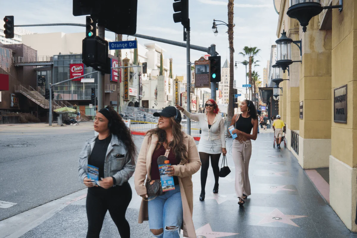 A group of tourists wearing headphones and holding brochures while strolling past sidewalk star plaques near the intersection of Hollywood Blvd and Orange Dr.