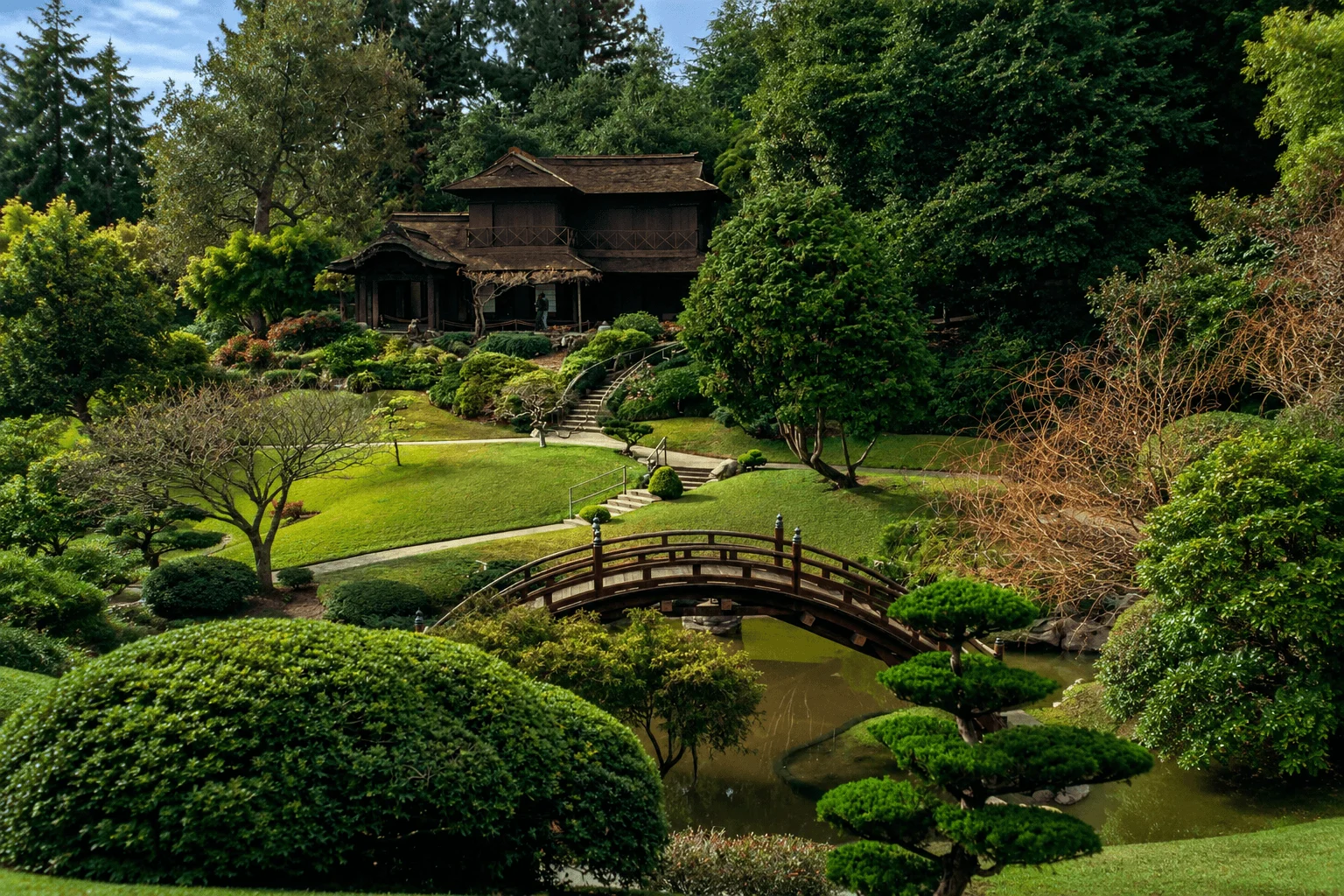 A lush, green Japanese garden featuring a traditional arched wooden footbridge over a quiet pond, winding stone pathways, manicured bushes, and a two-story wooden teahouse nestled in the trees.