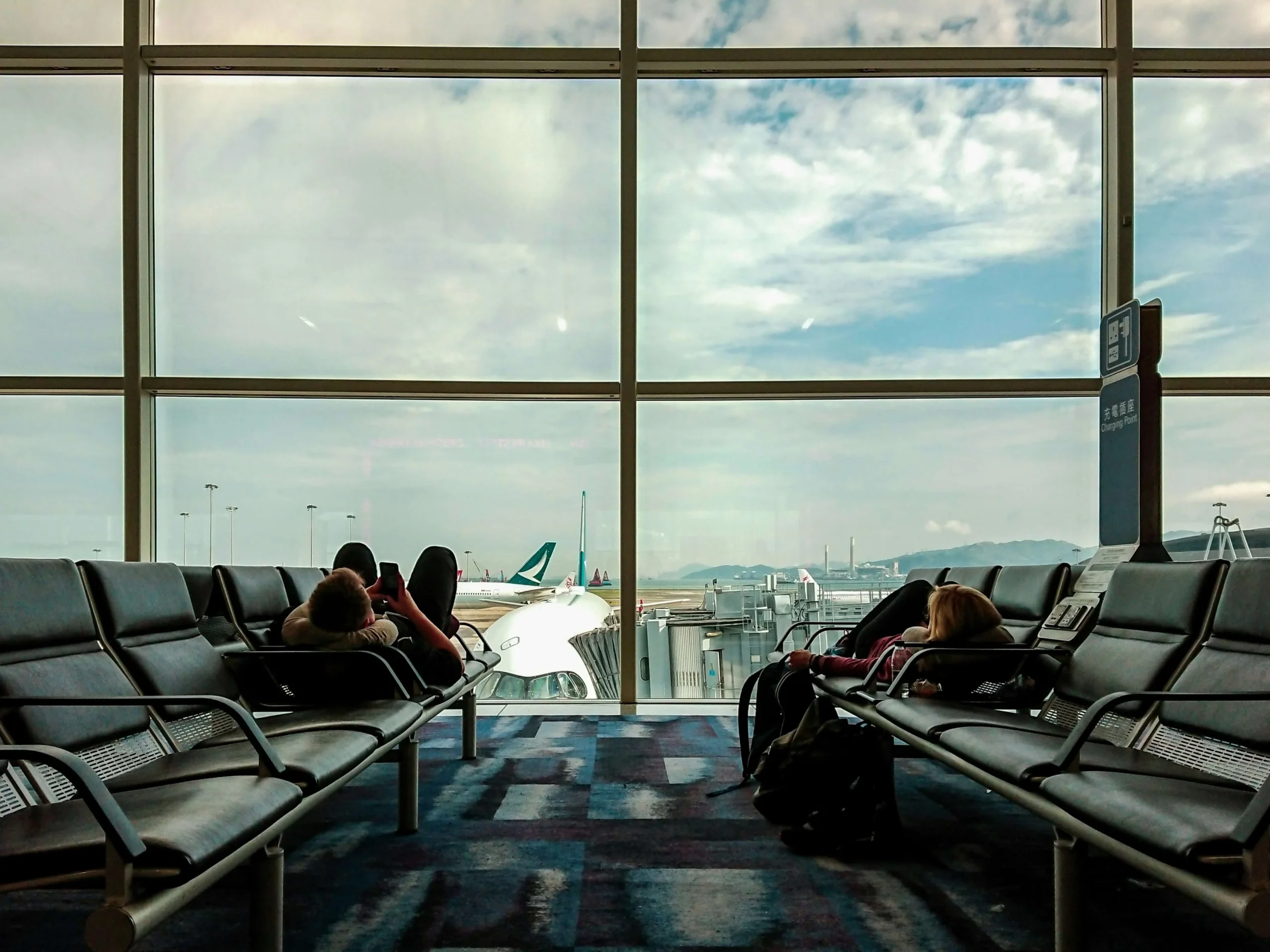 Two travelers lying down across black leather waiting area seats in an airport terminal, facing a giant glass wall with a view of parked airplanes on the tarmac.