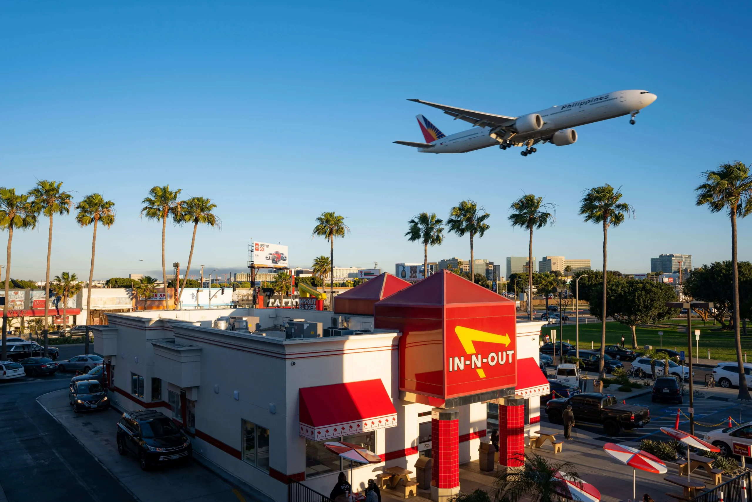High-angle view of the iconic In-N-Out Burger building near LAX surrounded by palm trees, with a commercial airplane flying low on final approach directly above it.