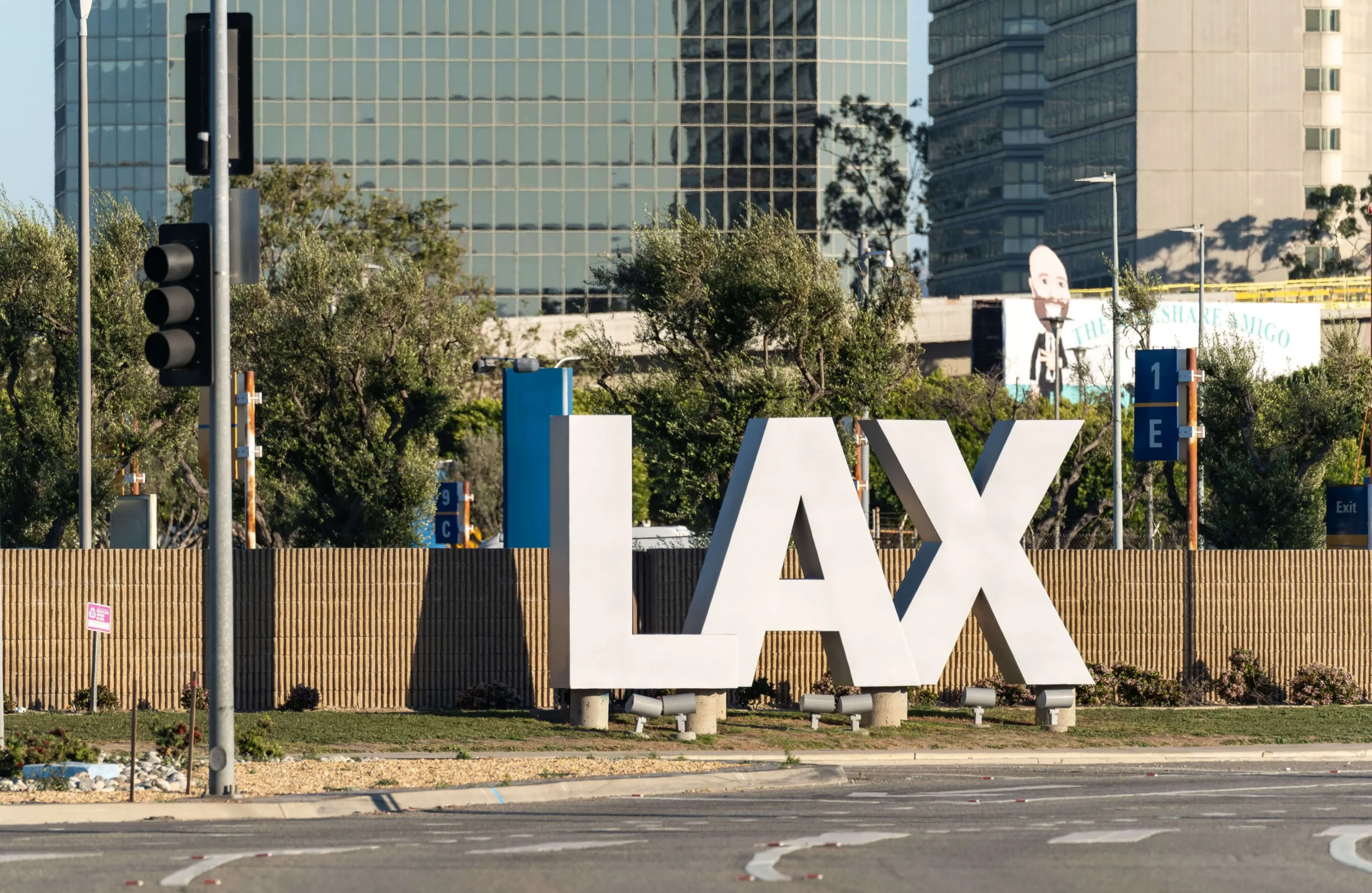 Large white block letters spelling out LAX along an entry road with modern high-rise office buildings and green trees in the background.