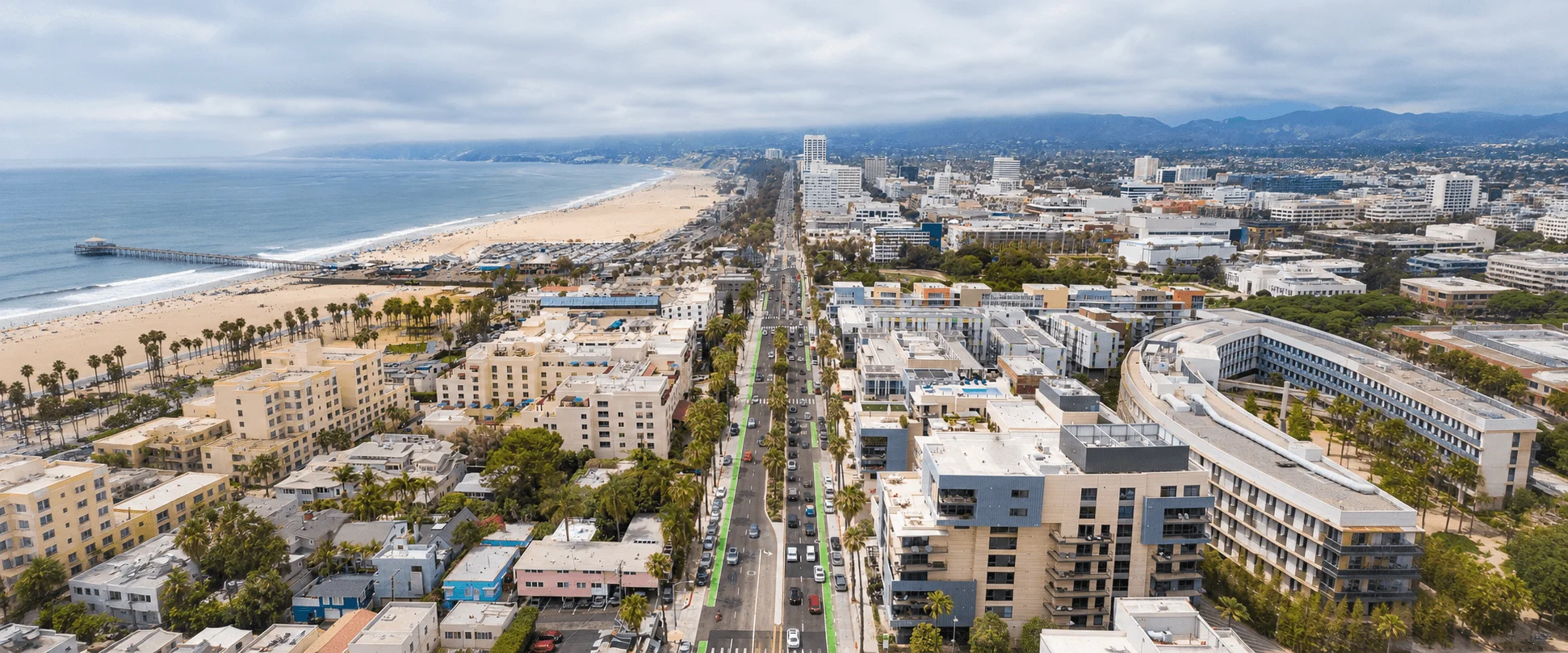 High-angle aerial view of the Santa Monica coastline showing the sandy beach, ocean pier, beachfront boulevard, and city buildings.