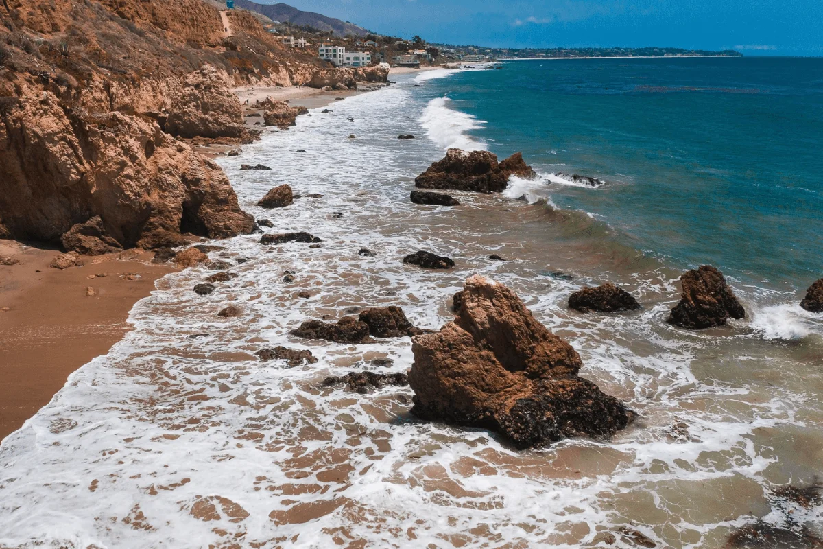 High-angle shot looking down at ocean waves crashing around prominent rock sea stacks along the sandy shoreline of El Matador Beach.