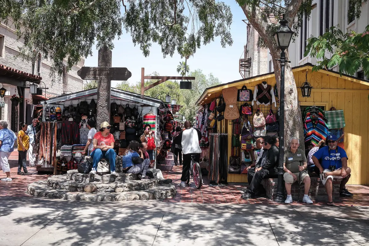 Tall carved wooden cross on a stone base surrounded by visitors and vendor stalls selling leather bags and serapes on Olvera Street