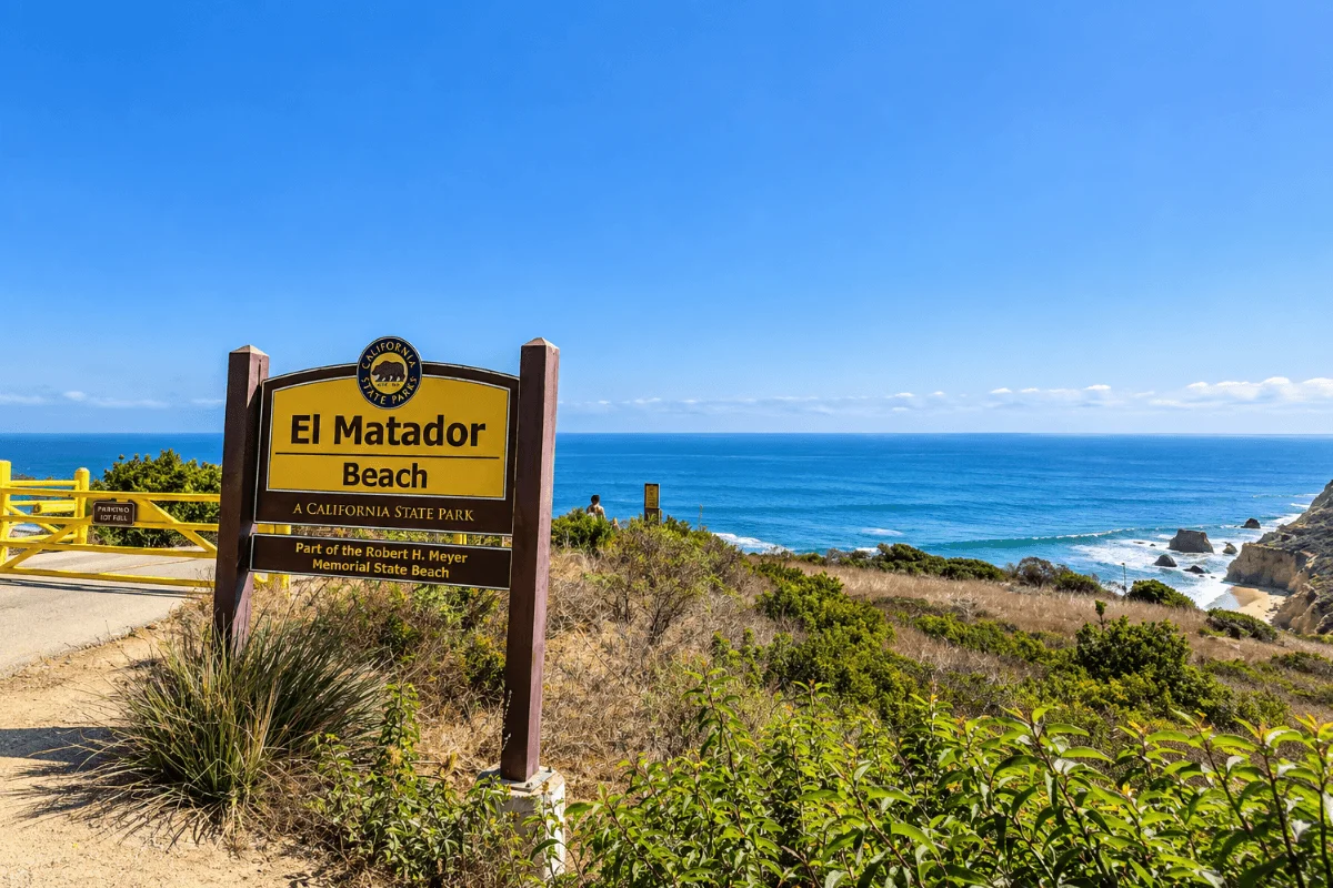 Official yellow California State Parks sign for El Matador Beach set against coastal vegetation with sweeping blue ocean views in Malibu.