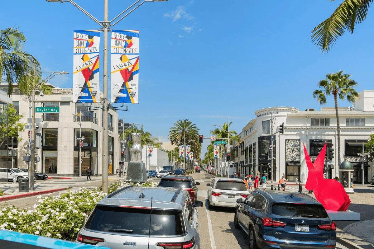 Traffic driving down Rodeo Drive past high-end luxury designer boutiques, palm trees, and a bright red origami rabbit sculpture.