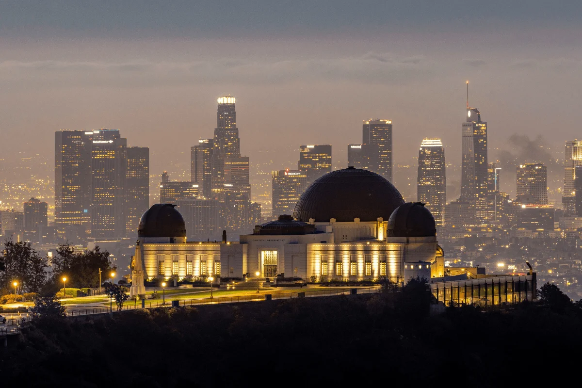 Illuminated Griffith Observatory at dusk with the glowing high-rise skyscrapers of Downtown Los Angeles in the background.