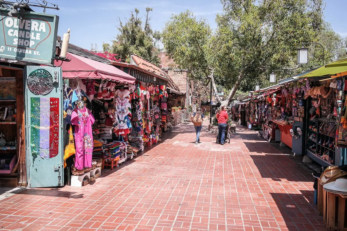 Olvera Candle Shop sign beside colorful market stalls selling Mexican dresses, blankets, and crafts along a sunny brick walkway